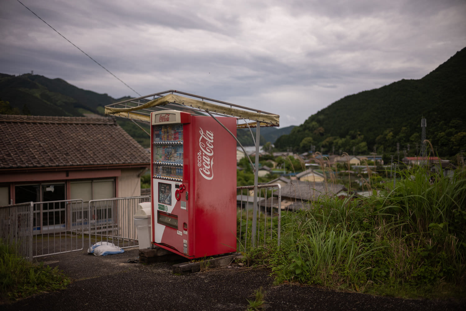 Consider the vending machine lonely on its hill, but, huh, look closely, much delights: Frame once holding up tarp to protect from the elements ripped away by decades of typhoons, now elegant (stitching, gently bent support poles, simple welded joints) crown of sadness for this unsung Hero of Thirst Relief, on side: Roman / Japanese syllabary mix of “ECOる” — roughly, “to Eco,” “to be Eco-friendly,” one assumes of the machine itself, algorithmic energy use for the benefit of earth, drinks cold but ethically cold — raised on concrete stilts to be perfectly even (straight bottle drops, no hangs), weeds to the right unkempt (but pleasant), but recycling pails (ECOる?) to left for cans and bottles in fine shape, sandbags holding half of small fence in place leading down to … a home? a shed? something uninhabited, and in the distance, the bay, small farming village of Mikisato flanked by mountain passes, unseen but heard: Colossal trucks rumbling back and forth carrying chopped wood and gravel from worksites up and down the coast.
